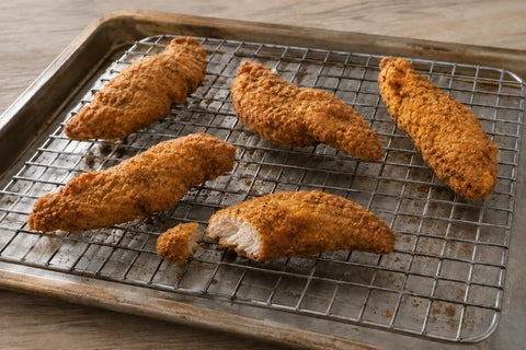 Golden breaded chicken tenderloins on a draining rack, with one cut to show the cooked interior.