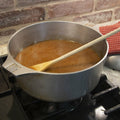 An aluminum stockpot with simmering brown roux and a wooden spoon sits on a black gas stovetop in a kitchen next to a red and white polka dot towel and brick backsplash