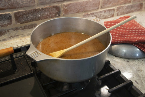 An aluminum stockpot with simmering brown roux and a wooden spoon sits on a black gas stovetop in a kitchen next to a red and white polka dot towel and brick backsplash