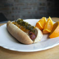 A hot dog with melted orange cheese and green relish on a white scalloped plate next to three orange wedges on a wooden table