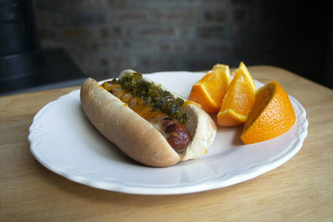 A hot dog with melted orange cheese and green relish on a white scalloped plate next to three orange wedges on a wooden table