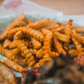 Close-up photograph of crispy crinkle-cut fries generously seasoned with reddish spice, piled in a white paper liner featuring a red logo, with a blurred dark food item in the foreground
