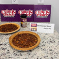 Two golden-brown pecan pies and a Cajun's Choice Breakfast Blend seasoning jar are showcased with three purple Cajun's Choice Premium Blend boxes and a white CC Breakfast Blend Pecan Pie label on a speckled granite kitchen countertop