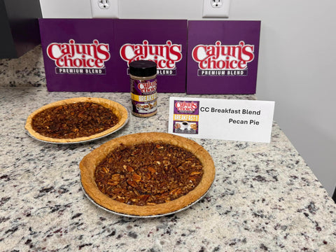 Two golden-brown pecan pies and a Cajun's Choice Breakfast Blend seasoning jar are showcased with three purple Cajun's Choice Premium Blend boxes and a white CC Breakfast Blend Pecan Pie label on a speckled granite kitchen countertop
