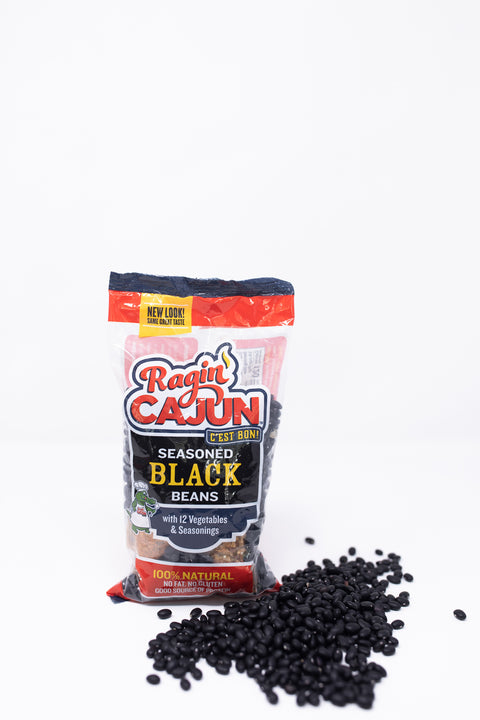 Product photography of Ragin' Cajun Seasoned Black Beans bag featuring a green alligator chef mascot and 'C'est Bon!' slogan, with black beans scattered on a white background