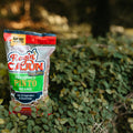 A Ragin' Cajun Seasoned Pinto Beans bag with its vibrant red and orange logo and "New Look!" banner, showing the speckled brown beans through its clear packaging, resting among lush green foliage outdoors