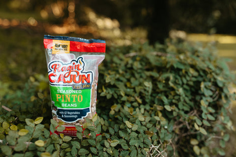 A Ragin' Cajun Seasoned Pinto Beans bag with its vibrant red and orange logo and "New Look!" banner, showing the speckled brown beans through its clear packaging, resting among lush green foliage outdoors