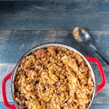 Photograph of a savory Jambalaya dish in a vibrant red enameled cast iron pot on a rustic wooden table featuring golden rice, sliced sausage, and shrimp alongside an ornate silver spoon