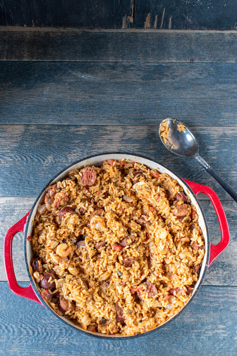 Photograph of a savory Jambalaya dish in a vibrant red enameled cast iron pot on a rustic wooden table featuring golden rice, sliced sausage, and shrimp alongside an ornate silver spoon