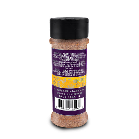 Product photography of an Acadian Kitchens spice bottle filled with brown seasoning, featuring a black flip-top lid and a vibrant purple label with white text detailing breakfast, snack, and dessert usage suggestions alongside yellow food icons on a white background