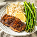 A close-up photograph of a healthy dinner plate featuring two spicy blackened salmon fillets, fluffy brown rice, bright green glistening asparagus, and a fresh lemon slice on a white textured plate