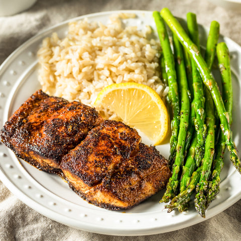 A close-up photograph of a healthy dinner plate featuring two spicy blackened salmon fillets, fluffy brown rice, bright green glistening asparagus, and a fresh lemon slice on a white textured plate