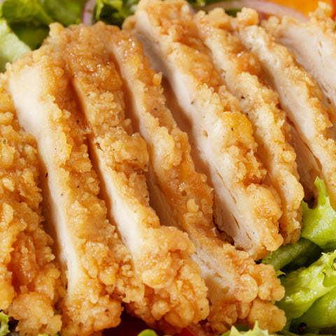A close-up photograph of crispy golden-brown sliced fried chicken breast on a bed of vibrant green lettuce with hints of red onion and orange tomato