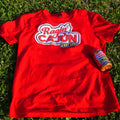 Photography of a vibrant red Ragin' Cajun t-shirt with a white and blue logo featuring 'C'est Bon' and a yellow chili pepper laid on bright green grass next to a matching red seasoning bottle under natural sunlight