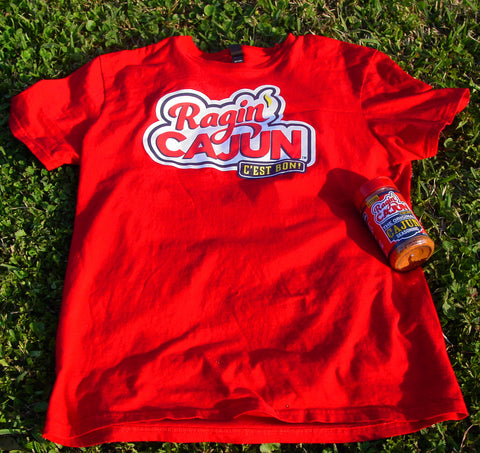 Photography of a vibrant red Ragin' Cajun t-shirt with a white and blue logo featuring 'C'est Bon' and a yellow chili pepper laid on bright green grass next to a matching red seasoning bottle under natural sunlight