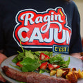 Photograph of a person wearing a black Ragin Cajun C'EST t-shirt holding a grey plate featuring a steak, roasted potatoes, and a green salad with red strawberries