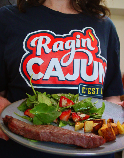 Photograph of a person wearing a black Ragin Cajun C'EST t-shirt holding a grey plate featuring a steak, roasted potatoes, and a green salad with red strawberries