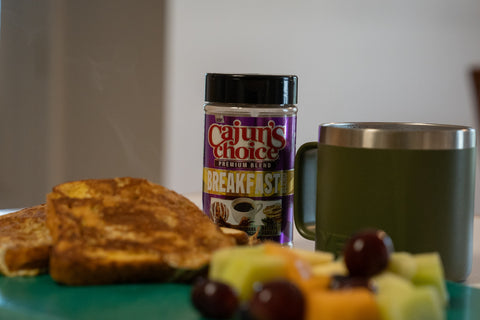A breakfast setting featuring a purple Cajun's Choice Premium Blend Breakfast seasoning container, golden French toast, an olive green Yeti mug, and a colorful fruit salad with red grapes and melon