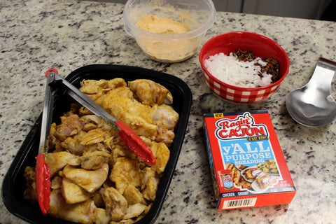 Photography of seasoned raw chicken pieces in a black tray with red tongs next to a Ragin' Cajun Y'all Purpose Breading box and a red and white checkered bowl of chopped onions and spices on a kitchen counter