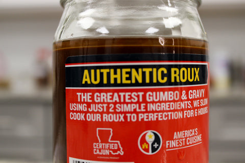 Photograph of a clear glass jar of dark brown "Authentic Roux" with a prominent red and black label featuring yellow "AUTHENTIC ROUX" text, "The Greatest Gumbo & Gravy" claim, and a "Certified Cajun" Louisiana outline logo