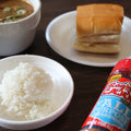 Photograph of a Cajun meal on a dark wooden table featuring a bowl of brown gumbo with green onions, a mound of fluffy white rice, a piece of golden bread, and a prominent red Ragin' Cajun seasoning bottle