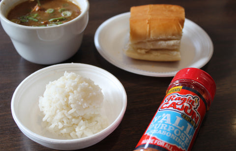 Photograph of a Cajun meal on a dark wooden table featuring a bowl of brown gumbo with green onions, a mound of fluffy white rice, a piece of golden bread, and a prominent red Ragin' Cajun seasoning bottle