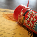 A Ragin' Cajun All Purpose Seasoning bottle with a bright red lid rests on a yellow restaurant menu displaying "Fried Catfish" and "Fried Oyster" entries, with orange spice powder spilled nearby