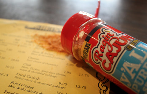 A Ragin' Cajun All Purpose Seasoning bottle with a bright red lid rests on a yellow restaurant menu displaying "Fried Catfish" and "Fried Oyster" entries, with orange spice powder spilled nearby