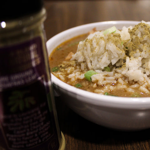 A white bowl of rich brown stew topped with fluffy white rice, golden seasoning, and vibrant green chopped scallions, presented on a dark wooden table with a blurred purple spice jar in the foreground