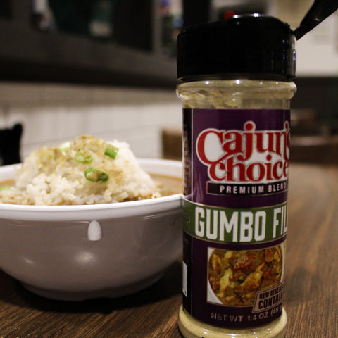 A "Cajun's Choice Gumbo Filé" seasoning bottle with a vibrant purple and green label stands beside a white bowl of gumbo topped with white rice and green onions on a wooden table