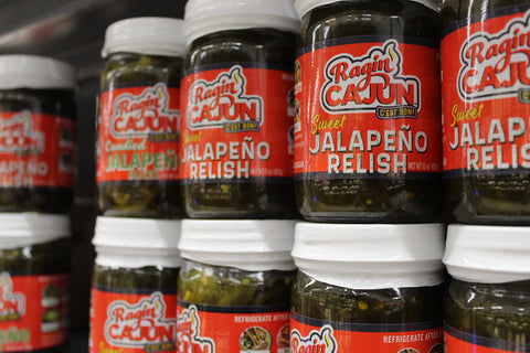 Photography of Ragin' Cajun Sweet Jalapeño Relish jars displayed on a store shelf, featuring vibrant red labels with 'C'est Bon' text, the green relish visible within, and white jar lids