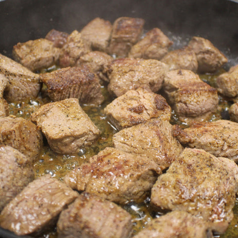 Photograph of seasoned beef chunks developing a golden brown crust while sizzling in hot oil within a dark frying pan