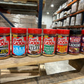Eight seasoning bottles from Ragin' Cajun and Cajun's Choice brands display bright red, black, and purple lids on a rustic wooden pallet shelf inside a warehouse stocked with many packaged goods