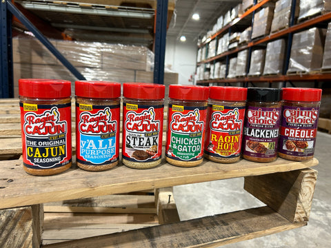 Eight seasoning bottles from Ragin' Cajun and Cajun's Choice brands display bright red, black, and purple lids on a rustic wooden pallet shelf inside a warehouse stocked with many packaged goods