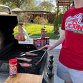 A person wearing a red "Ragin' Cajun C'est Bon!" t-shirt grills three steaks on a black Char-Broil gas grill on an outdoor patio, surrounded by several Ragin' Cajun seasoning bottles and a pineapple-shaped terracotta planter