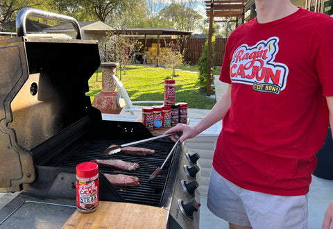 A person wearing a red "Ragin' Cajun C'est Bon!" t-shirt grills three steaks on a black Char-Broil gas grill on an outdoor patio, surrounded by several Ragin' Cajun seasoning bottles and a pineapple-shaped terracotta planter