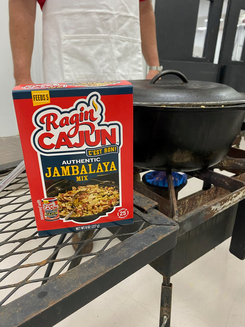 Photograph of a red Ragin' Cajun Authentic Jambalaya Mix box next to a black cast iron pot simmering on a rusty outdoor burner with a bright blue flame, a person in a white apron visible behind