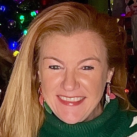 Woman with blonde hair and a green sweater in front of a decorated Christmas tree.