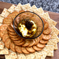 A creamy cheese ball topped with dark green relish in a clear glass bowl is surrounded by an array of golden round crackers and square saltine crackers artfully arranged on a rustic wooden charcuterie board set against a speckled grey granite countertop
