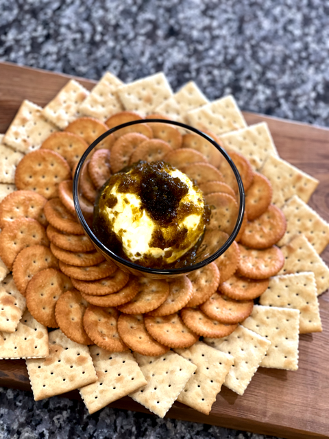 A creamy cheese ball topped with dark green relish in a clear glass bowl is surrounded by an array of golden round crackers and square saltine crackers artfully arranged on a rustic wooden charcuterie board set against a speckled grey granite countertop