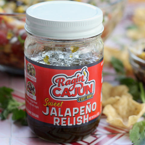 A clear glass jar of Ragin' Cajun Sweet Jalapeño Relish with a vibrant red and white label displaying a flame logo, filled with green relish showing seeds and condensation, topped with a white metal lid, set against a blurred background of fresh cilantro and tortilla chips on a striped surface