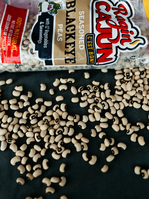 A clear bag of Ragin' Cajun Seasoned Blackeye Peas featuring a vibrant red brand logo and cartoon alligator chef, with many beige blackeye peas with black "eyes" scattered across a dark surface, highlighting "C'EST BON!" and "100% NATURAL" labels