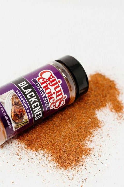 Product photography of a purple Cajun's Choice Blackened Seasoning & Rub bottle on a white surface with spilled reddish-brown seasoning and a label showing blackened meat