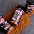 Photograph of three purple-labeled Cajun's Choice Premium Blend Blackened Seasoning & Rub bottles on a dark gray surface with vibrant reddish-orange spice scattered around, each label featuring an image of blackened meat on a white plate with green garnish