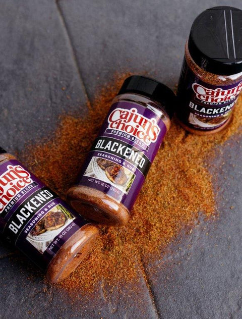 Photograph of three purple-labeled Cajun's Choice Premium Blend Blackened Seasoning & Rub bottles on a dark gray surface with vibrant reddish-orange spice scattered around, each label featuring an image of blackened meat on a white plate with green garnish