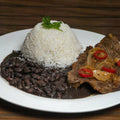 A Brazilian-style meal on a white plate featuring a mound of white rice with a fresh parsley sprig, dark black beans, and a steak topped with golden caramelized onions and bright red pepper rings on a wooden surface