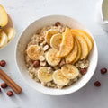 Photograph of a creamy oatmeal bowl generously topped with sliced bananas, fanned yellow apple slices, sprinkled cinnamon, and whole hazelnuts, artfully arranged with a side of apple and banana slices, cinnamon sticks, and a white milk pitcher on a bright textured countertop