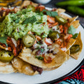 Photograph of a white plate piled high with loaded nachos topped with vibrant green guacamole, bright jalapeño slices, and gooey melted cheese, set on a dark wooden table next to a colorful patterned textile
