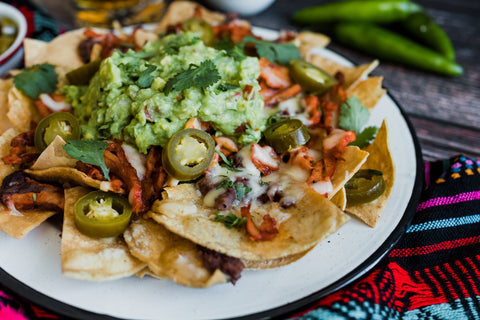 Photograph of a white plate piled high with loaded nachos topped with vibrant green guacamole, bright jalapeño slices, and gooey melted cheese, set on a dark wooden table next to a colorful patterned textile