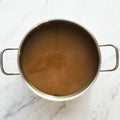 Overhead photograph of a stainless steel pot containing rich brown broth resting on a white marble surface with subtle grey veining